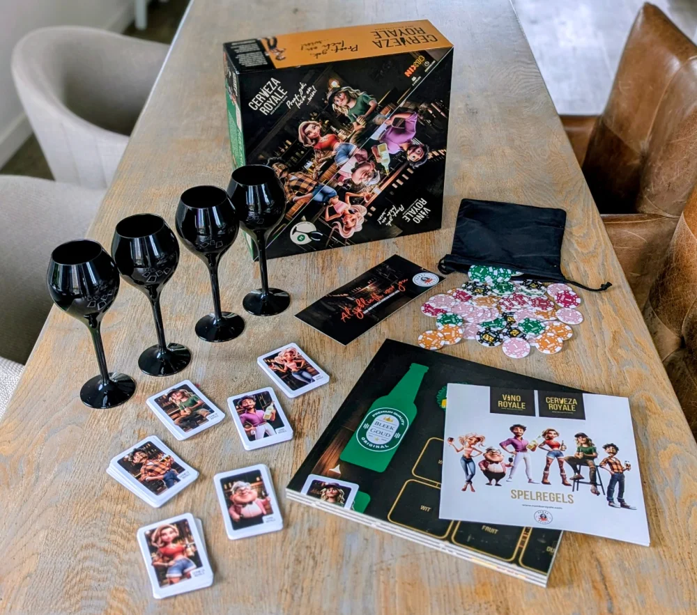 Ron van Noord — Visual design, Grafisch ontwerp, DTP, Visual design — Vino & Cerveza Royale — Beeld: Overzicht van de speldoos, zwarte proefglazen, karakterkaarten, roulettechips, spelborden en spelregels van Vino & Cerveza Royale op een houten tafel. — Project: Een spel vol smaak, spanning en karakter.